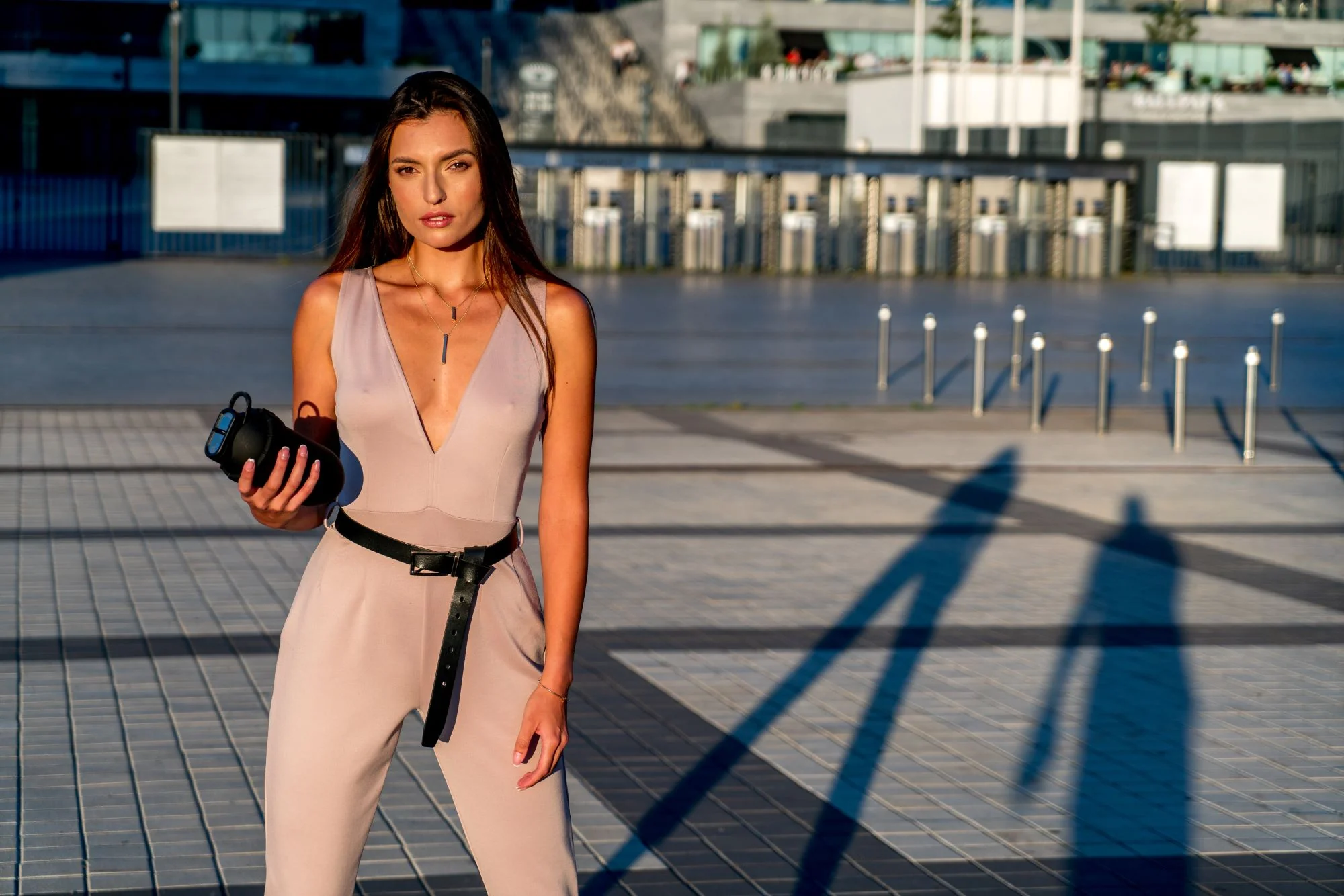 Woman in a beige jumpsuit holding a black object, standing outdoors with long shadows.