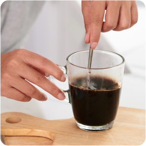 Person stirring black coffee in a clear mug on a wooden board.