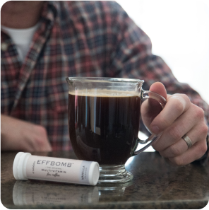A person holding a glass mug of coffee beside an EFFBOMB multivitamin container.