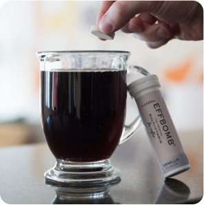 A hand dropping a vanilla-flavored tablet into a glass mug of black coffee.