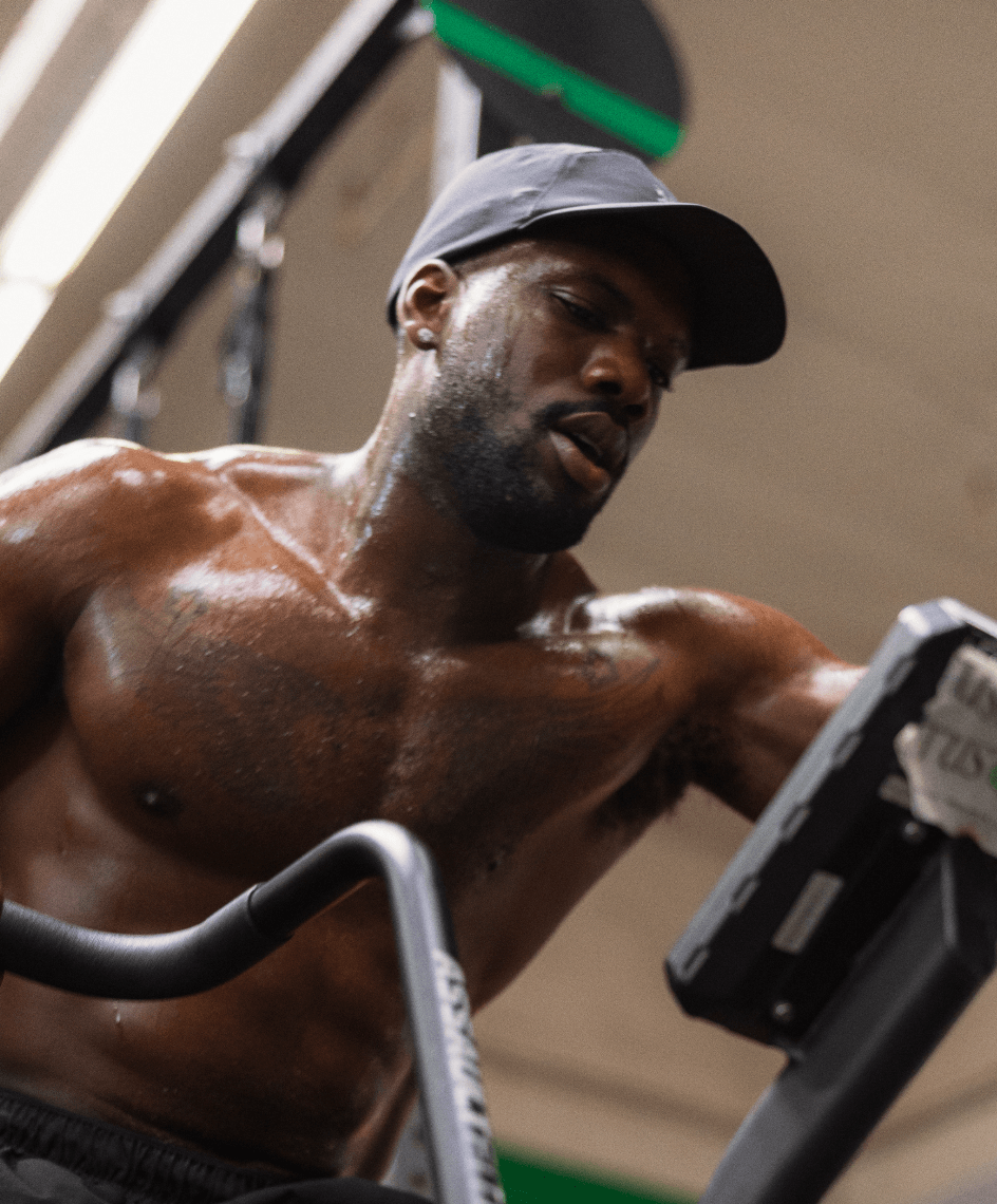 A sweaty, muscular man in a black cap works out on an exercise machine at a gym.