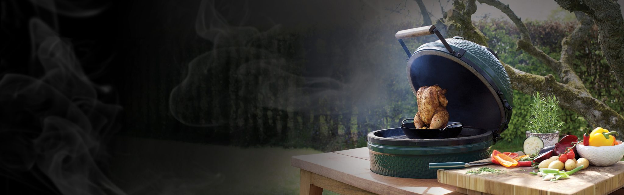 A whole chicken cooking in a green outdoor smoker grill next to a cutting board with fresh vegetables.