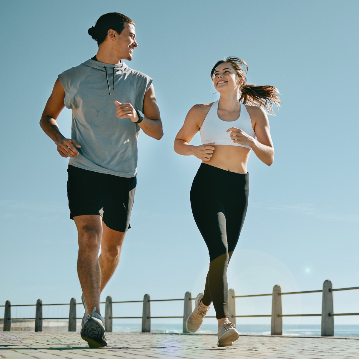 Two people jogging outdoors on a sunny day, smiling at each other.