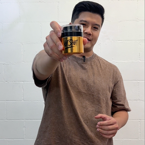Person holding a small gold container labeled 'Hot Shot' against a white brick wall.