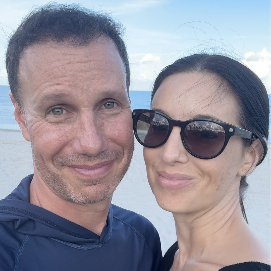A couple smiling on a beach with the ocean in the background.