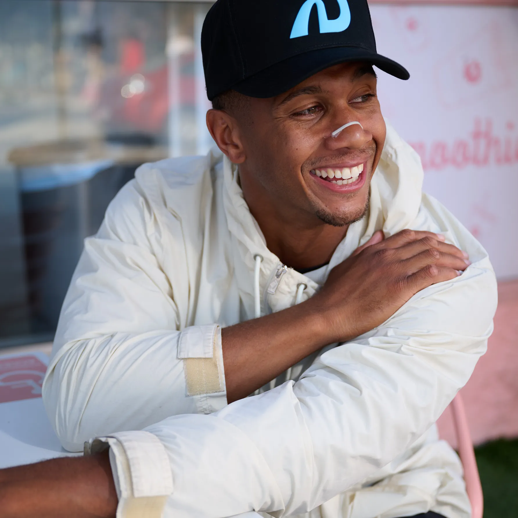 A smiling man in a white jacket, black cap, and nasal strip sits at a table.