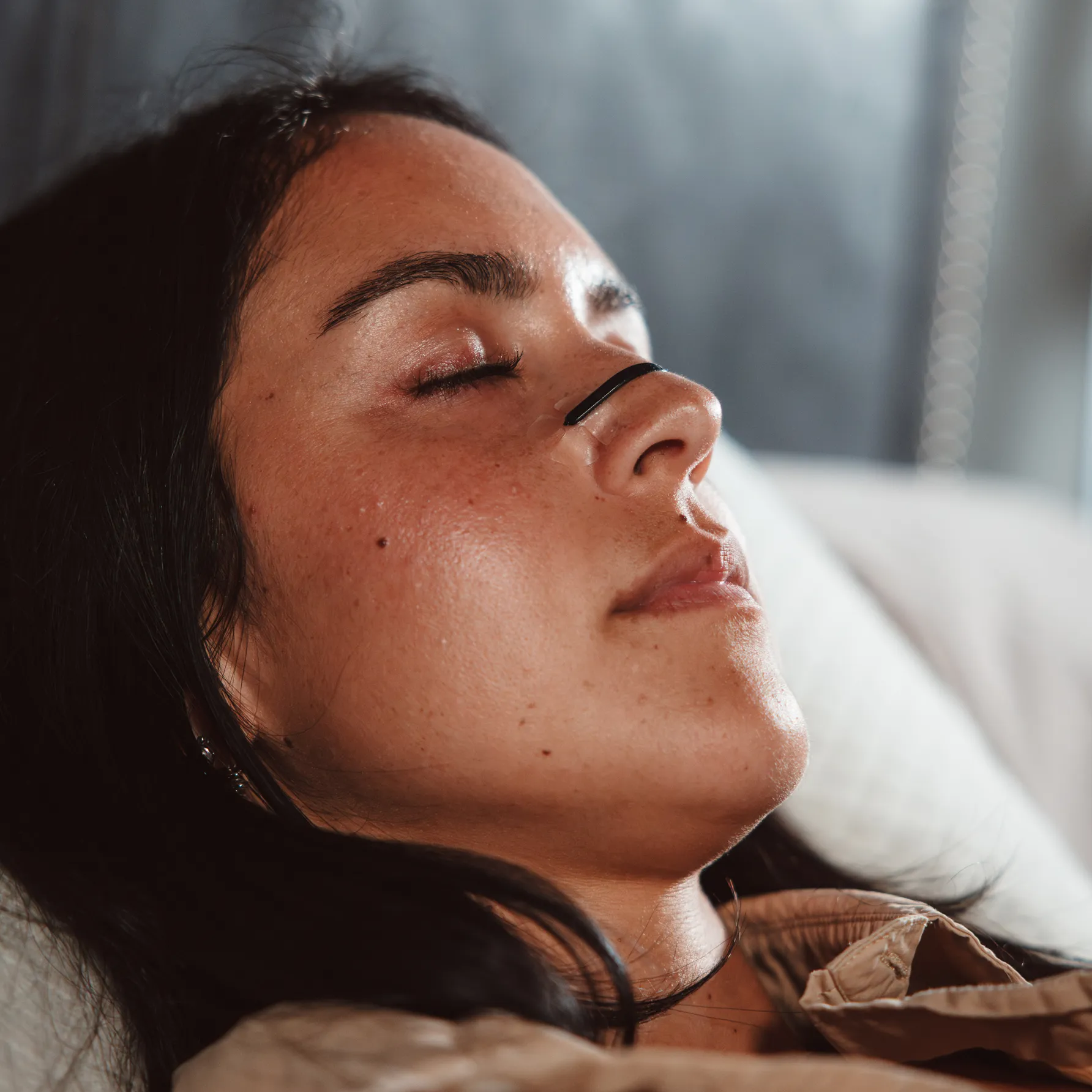 A close-up of a woman resting with her eyes closed, wearing a nasal strip across her nose.