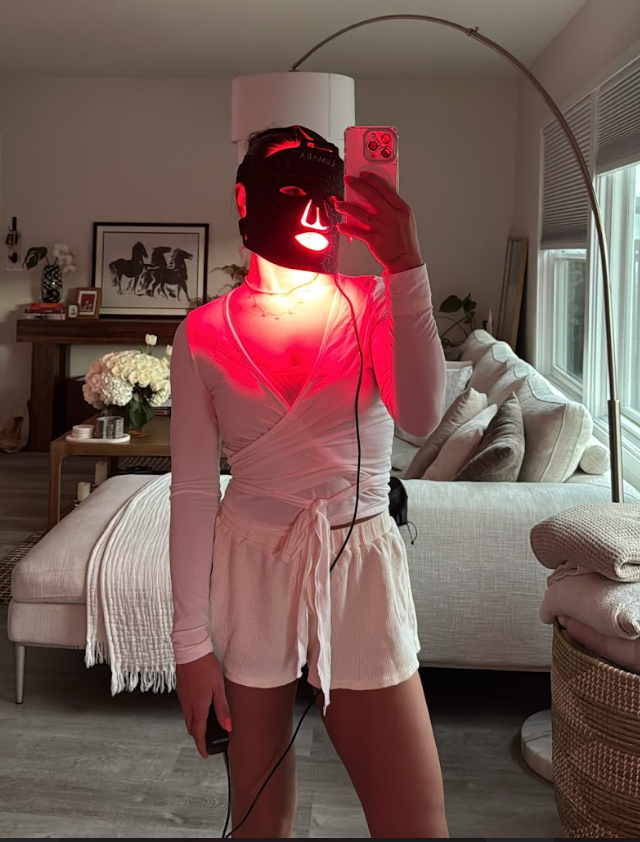 Person wearing a red LED face mask, taking a selfie in a living room.