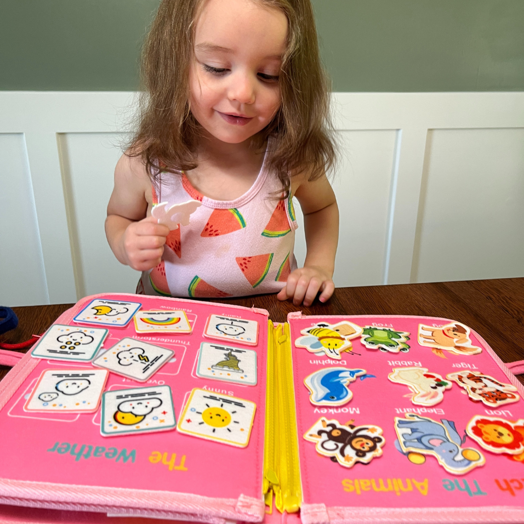 Child playing with an educational book featuring animal and weather illustrations on a pink background.
