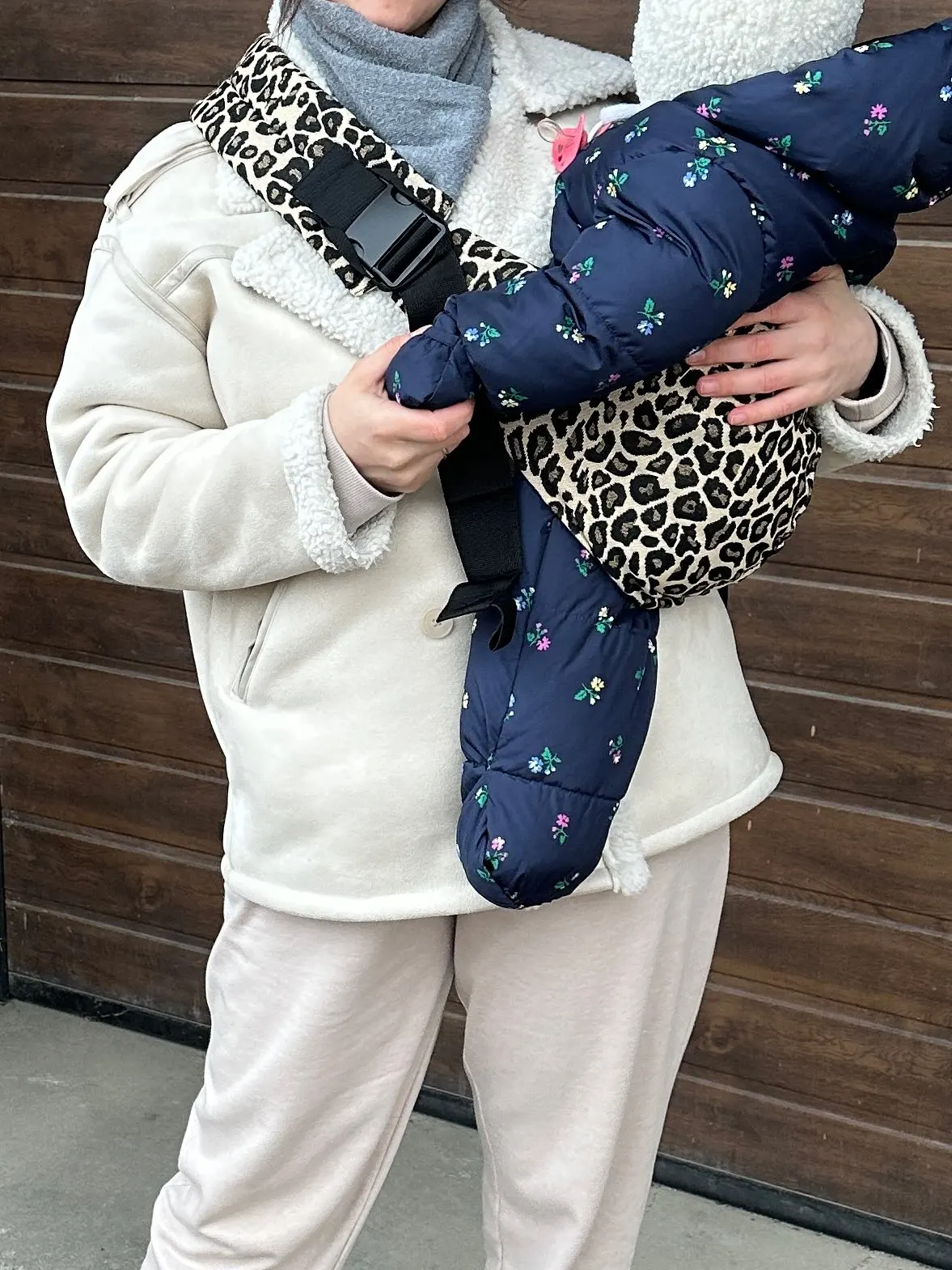 Person holding a baby in a floral snowsuit and leopard print carrier.