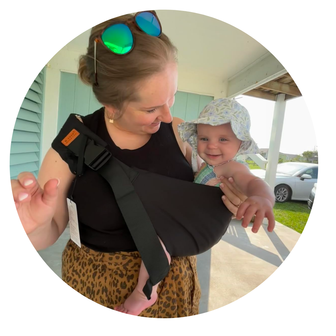 Woman holding a smiling baby in a sling, both outdoors.