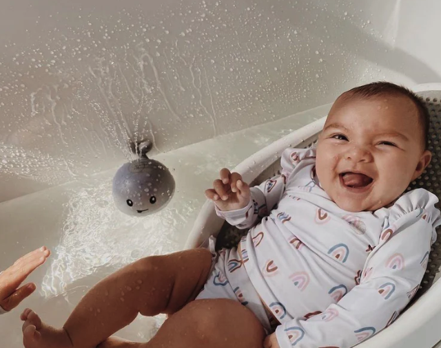 A laughing baby in a rainbow swimsuit sits in a bath seat next to a spraying whale toy.