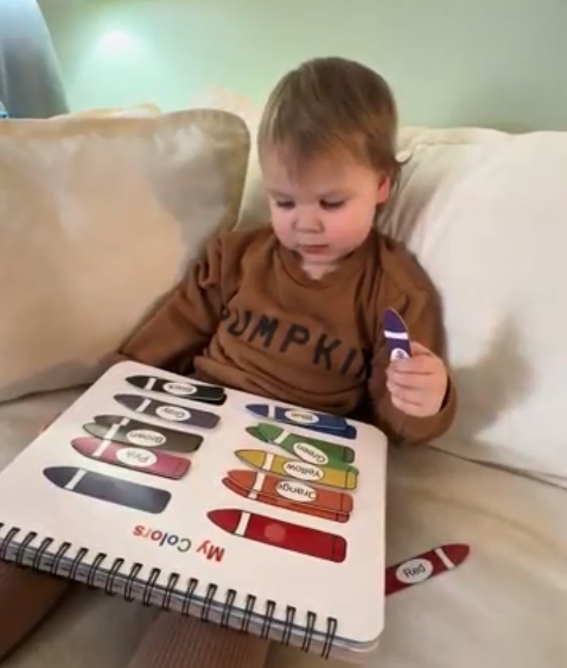 A toddler sits on a couch with a 'My Colors' puzzle book, holding a purple crayon piece.