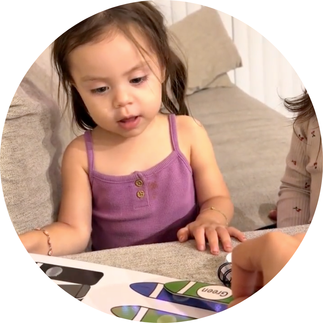 A young girl in a purple top looks at a color-matching book with crayon-shaped pieces.