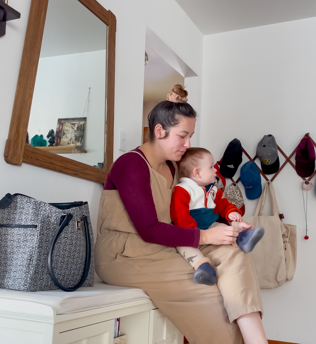 A woman sitting with a baby on a bench, with hats hanging on the wall.