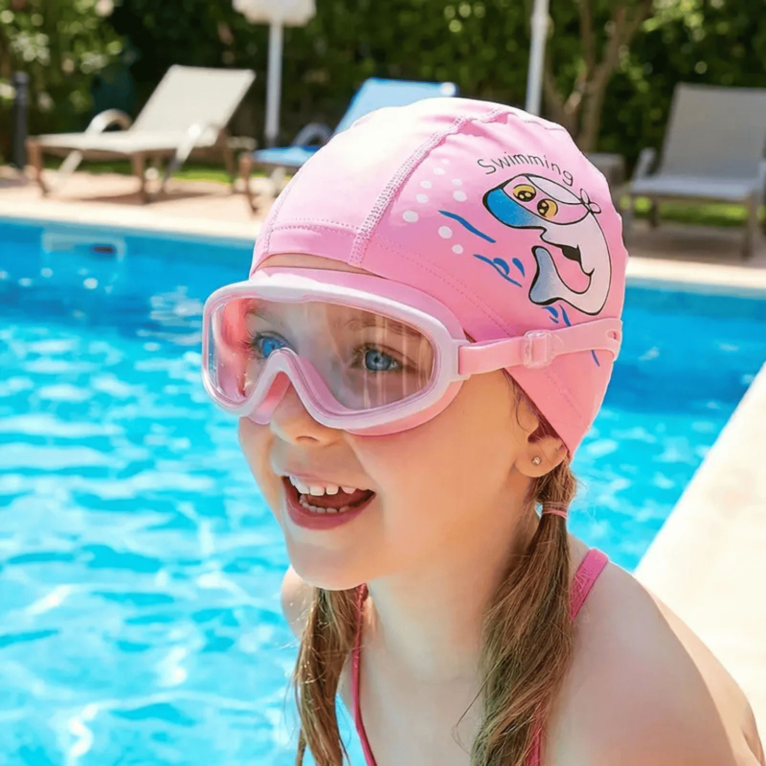 A smiling young girl in a pink swim cap and goggles sits by the edge of a pool.