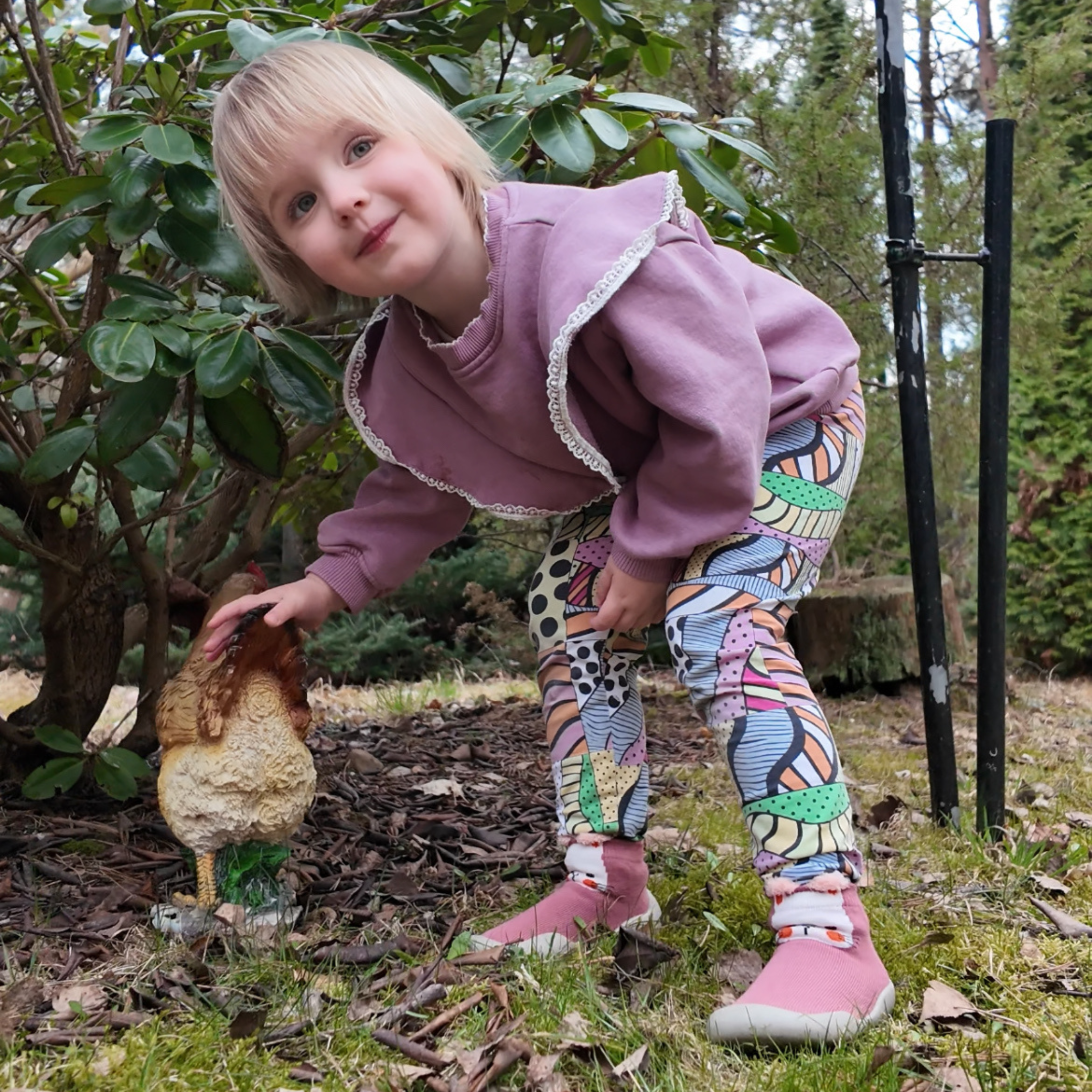 Child in colorful pants touching a garden rooster statue.