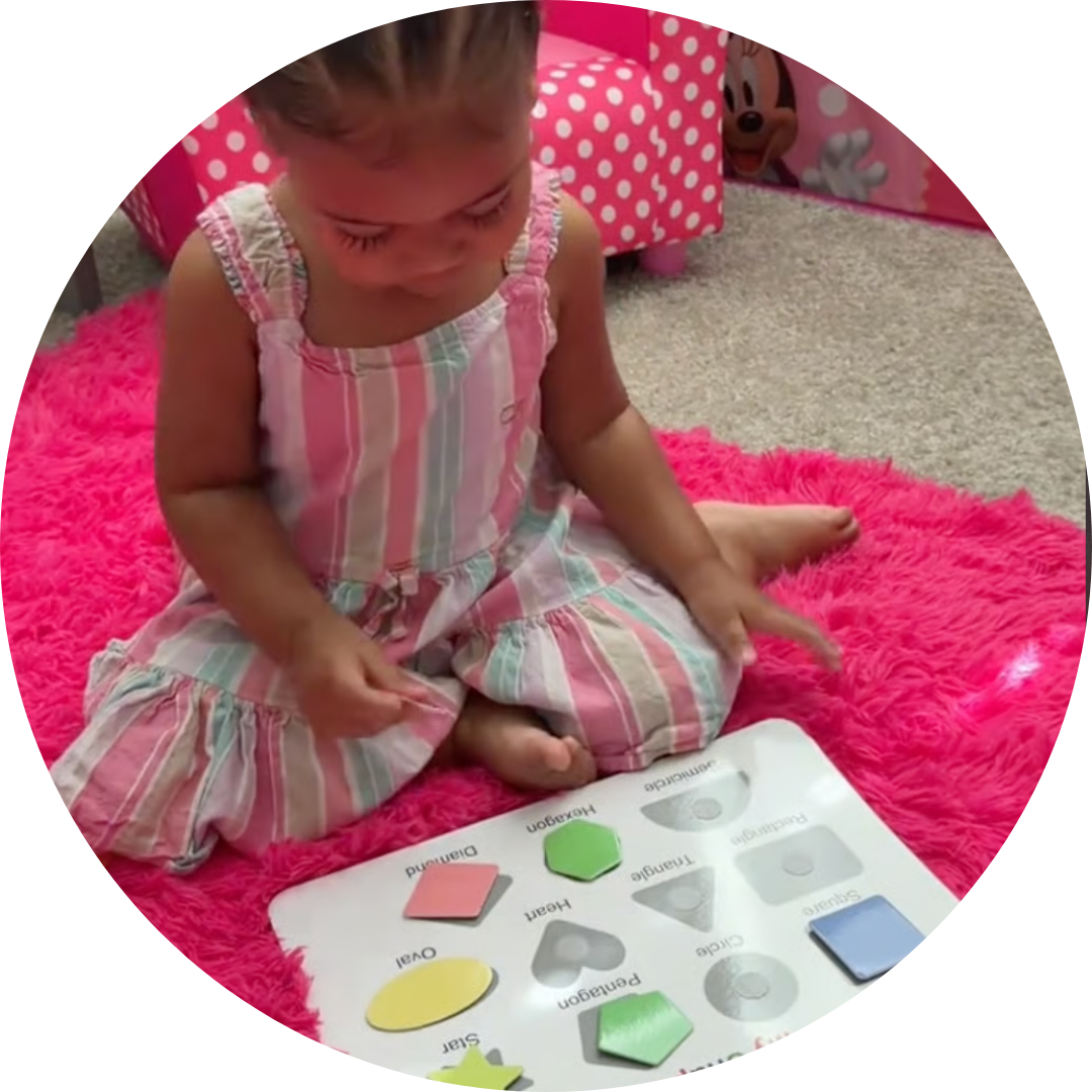 A young girl sits on a fluffy pink rug, playing with a wooden shape puzzle.