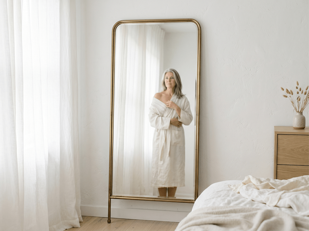 A woman in a bathrobe looks at her reflection in a full-length mirror in a bedroom.