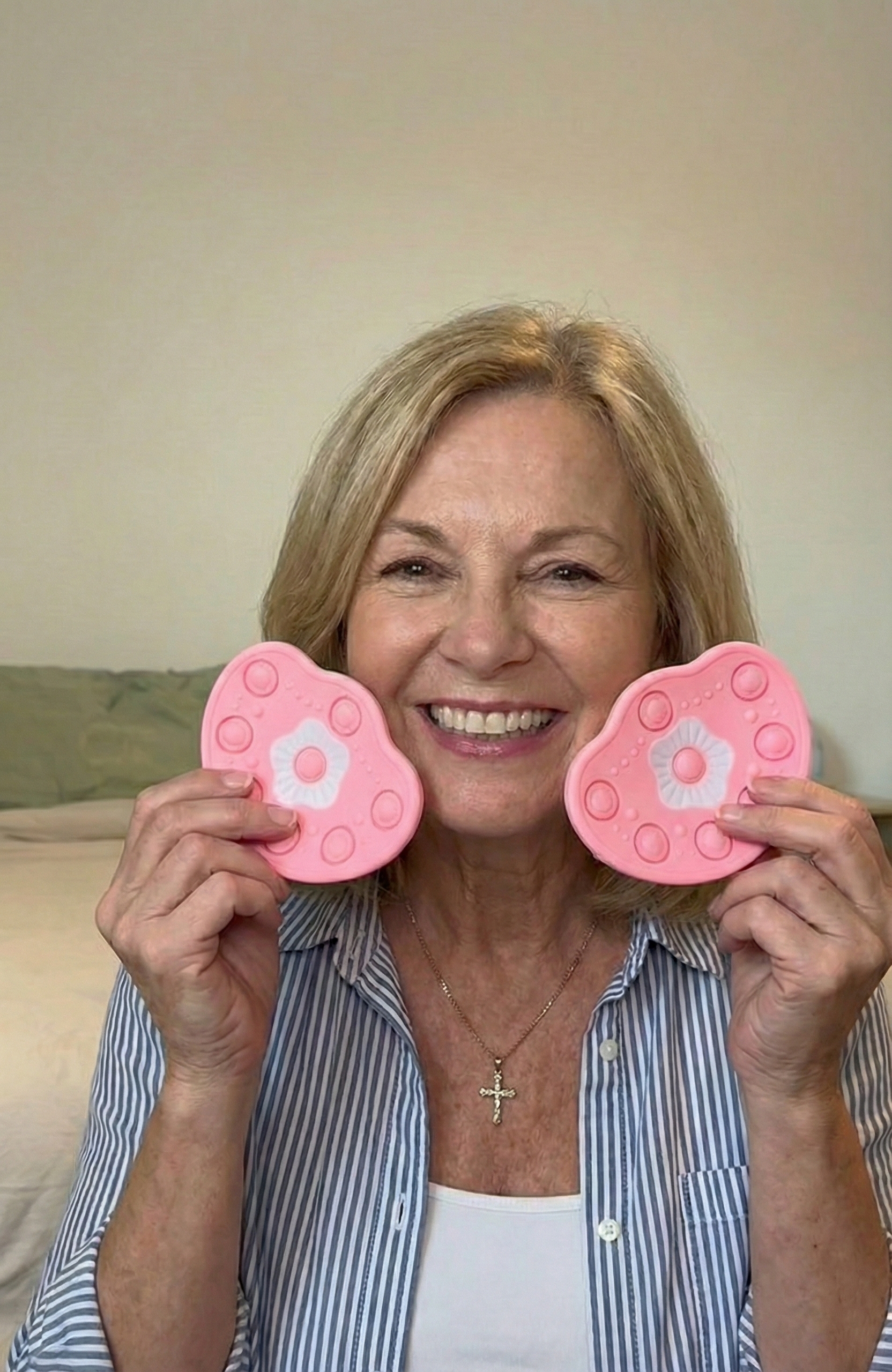A smiling woman holds two pink, flower-shaped fidget toys up to her cheeks in a bedroom.