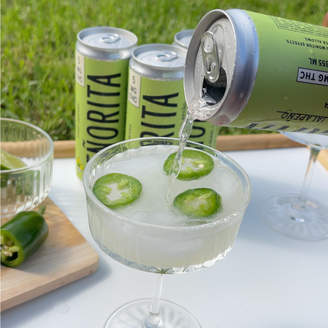Beverage being poured into a glass with jalapeño slices, next to cans and a cutting board with jalapeños.