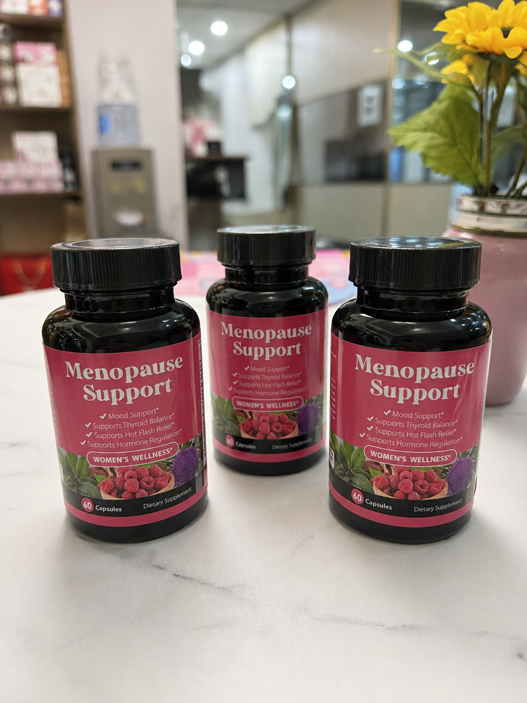 Three bottles of 'Menopause Support' dietary supplement on a white table next to a vase of yellow flowers.