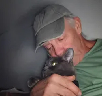 A man in a baseball cap closes his eyes while hugging a grey cat.