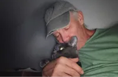 A man in a grey cap holds a grey cat close and kisses its head.