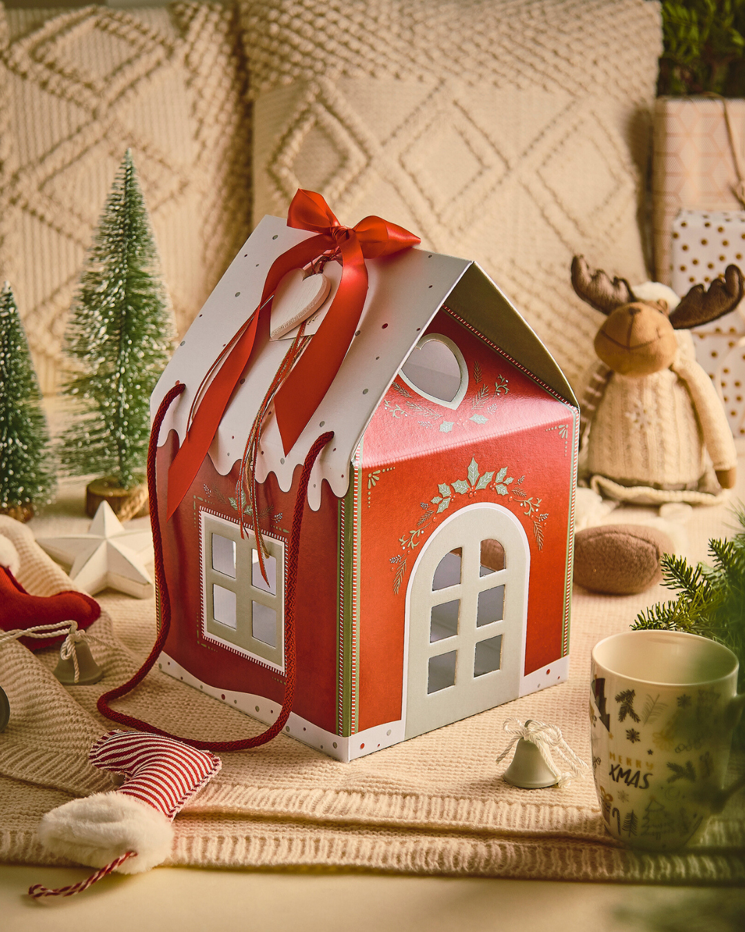 Gingerbread house gift box with red ribbon, surrounded by holiday decor and pine trees.
