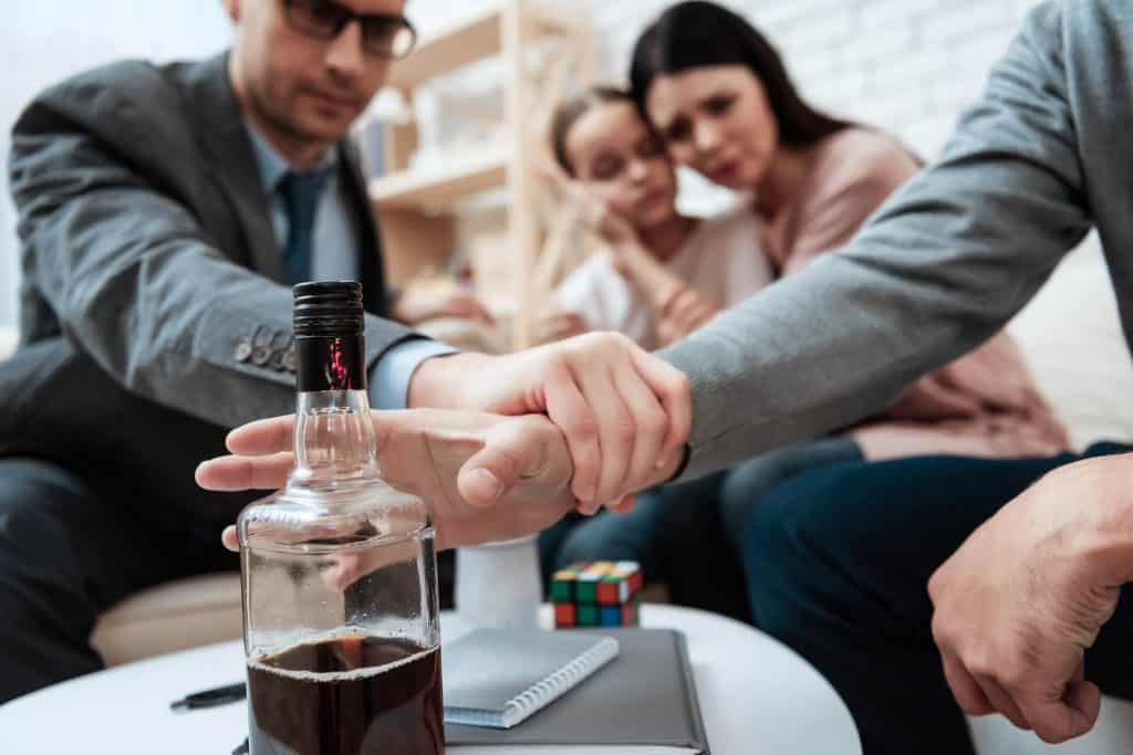 Person preventing another from taking a bottle of alcohol, with concerned people in the background.