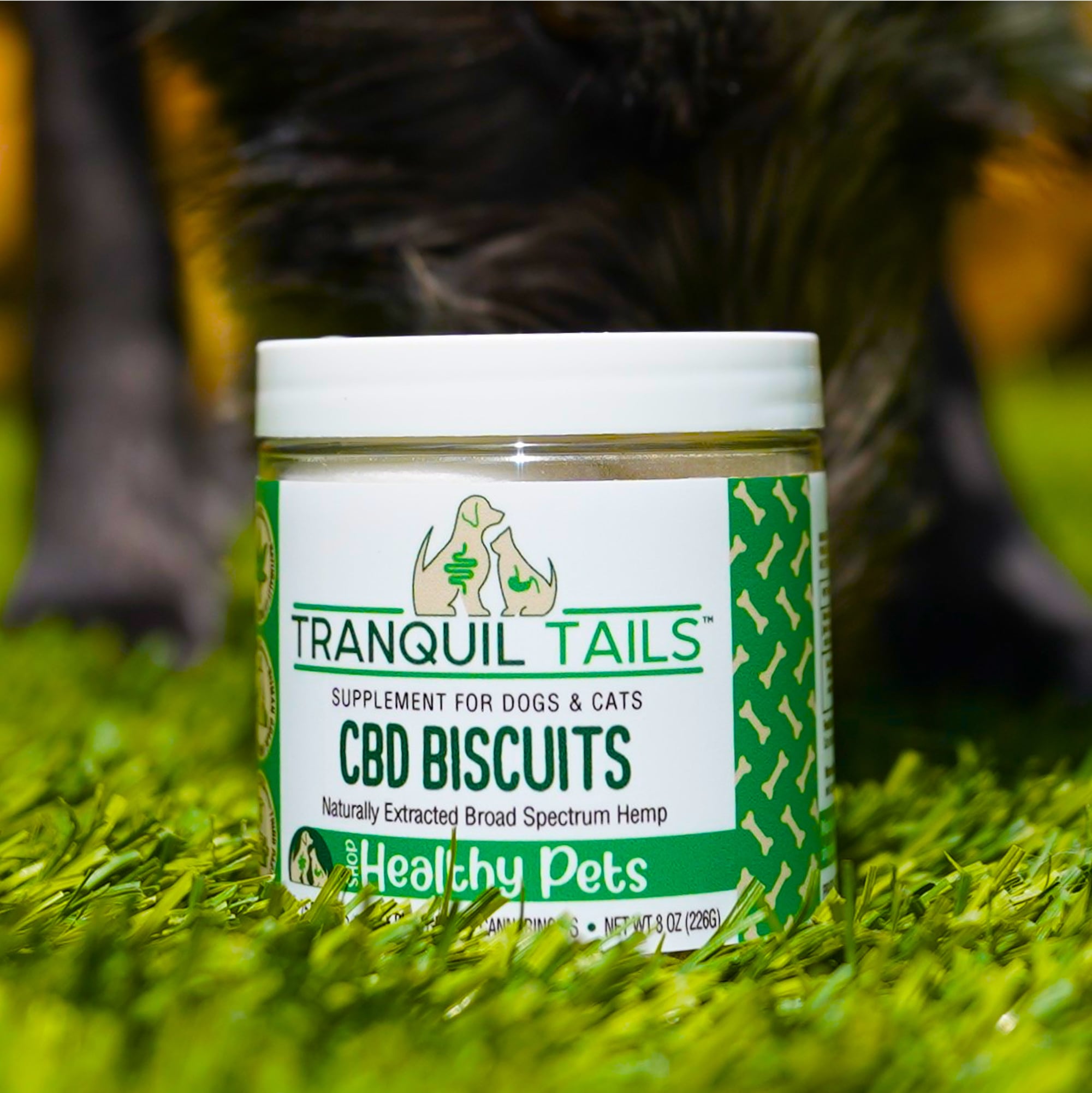 Jar of CBD biscuits for dogs and cats on grass with animal legs behind.