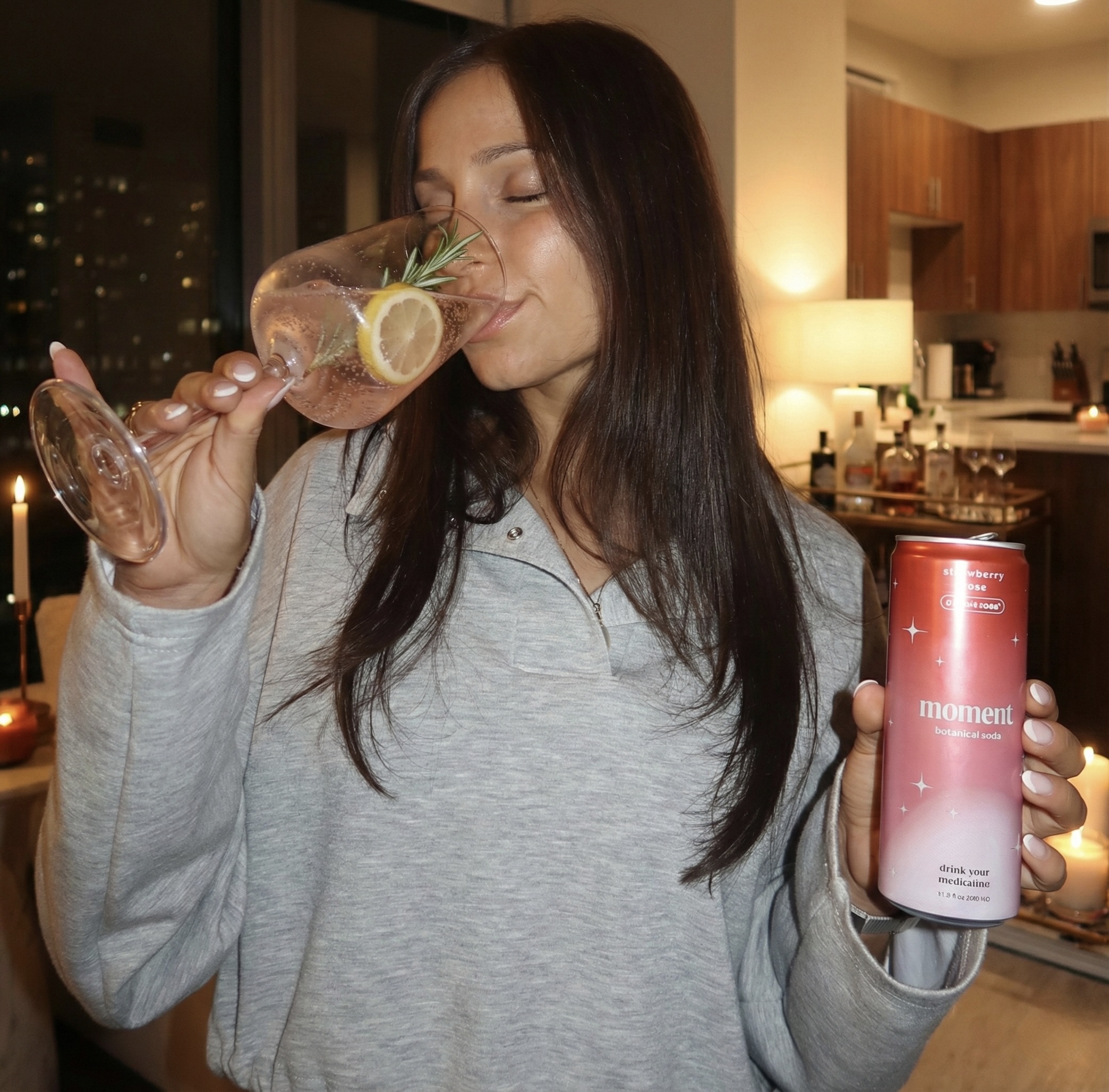 Person sipping from a glass with lemon and holding a can in a cozy kitchen.