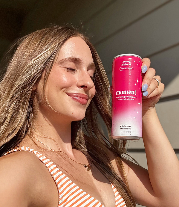 A woman with her eyes closed smiles while holding a pink can of Moment sparkling botanical drink.