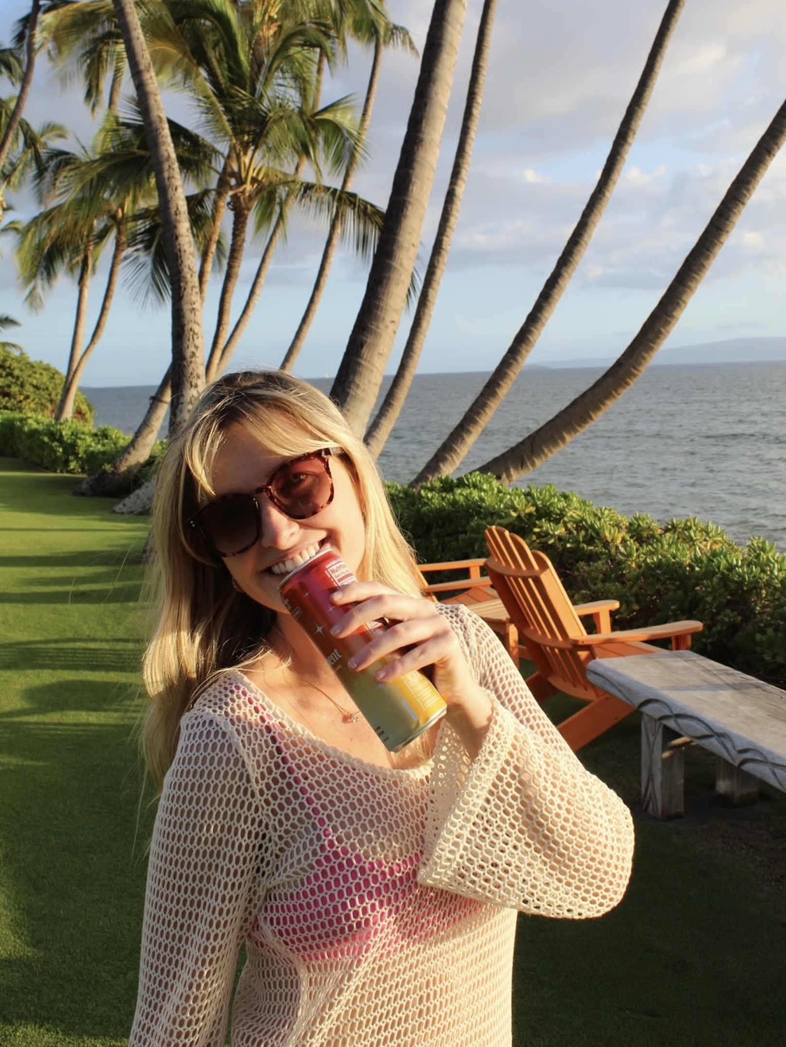 Person in sunglasses holding a can, standing near palm trees by the ocean.