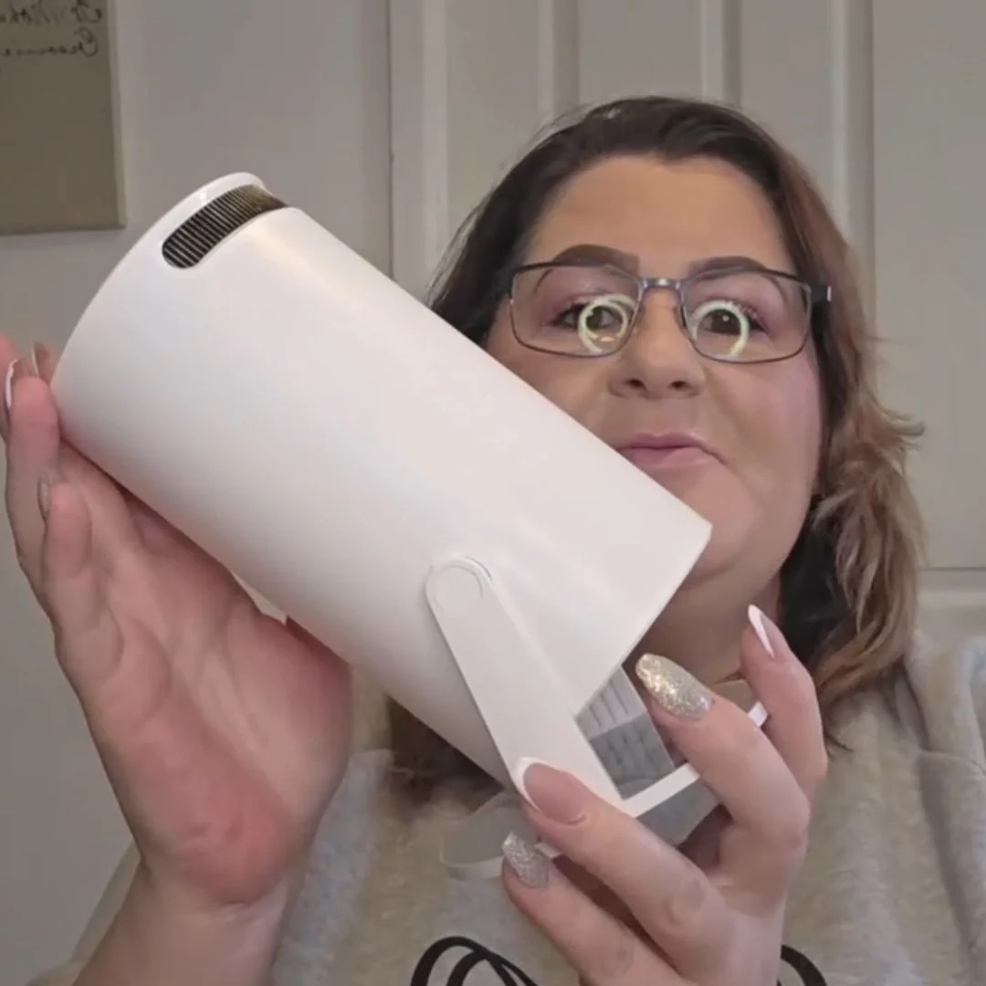 A woman with glasses holds up a white, cylindrical portable projector, with a ring light reflected in her glasses.