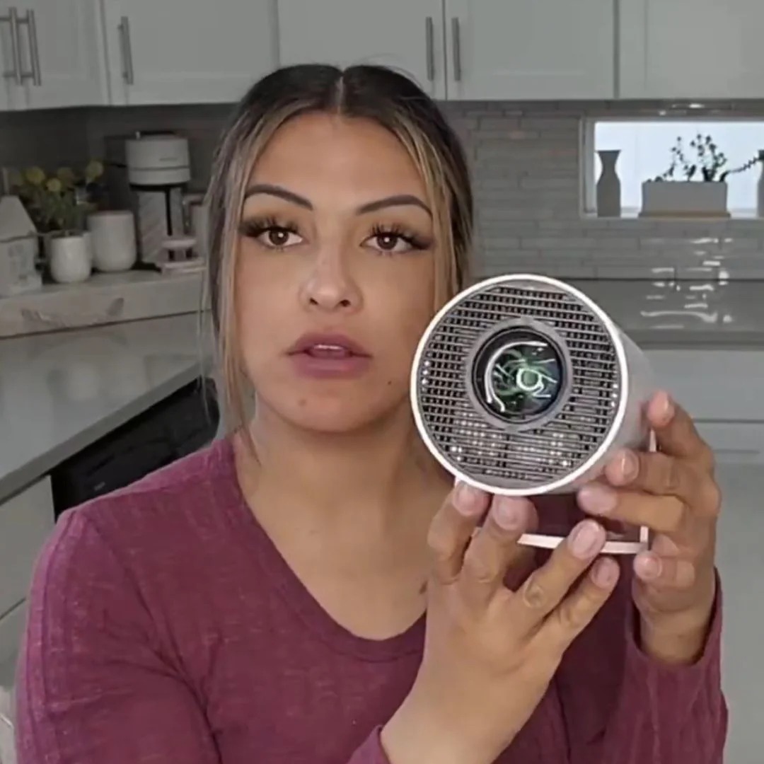 A woman in a kitchen holds up a small, white, cylindrical projector with a grated front.