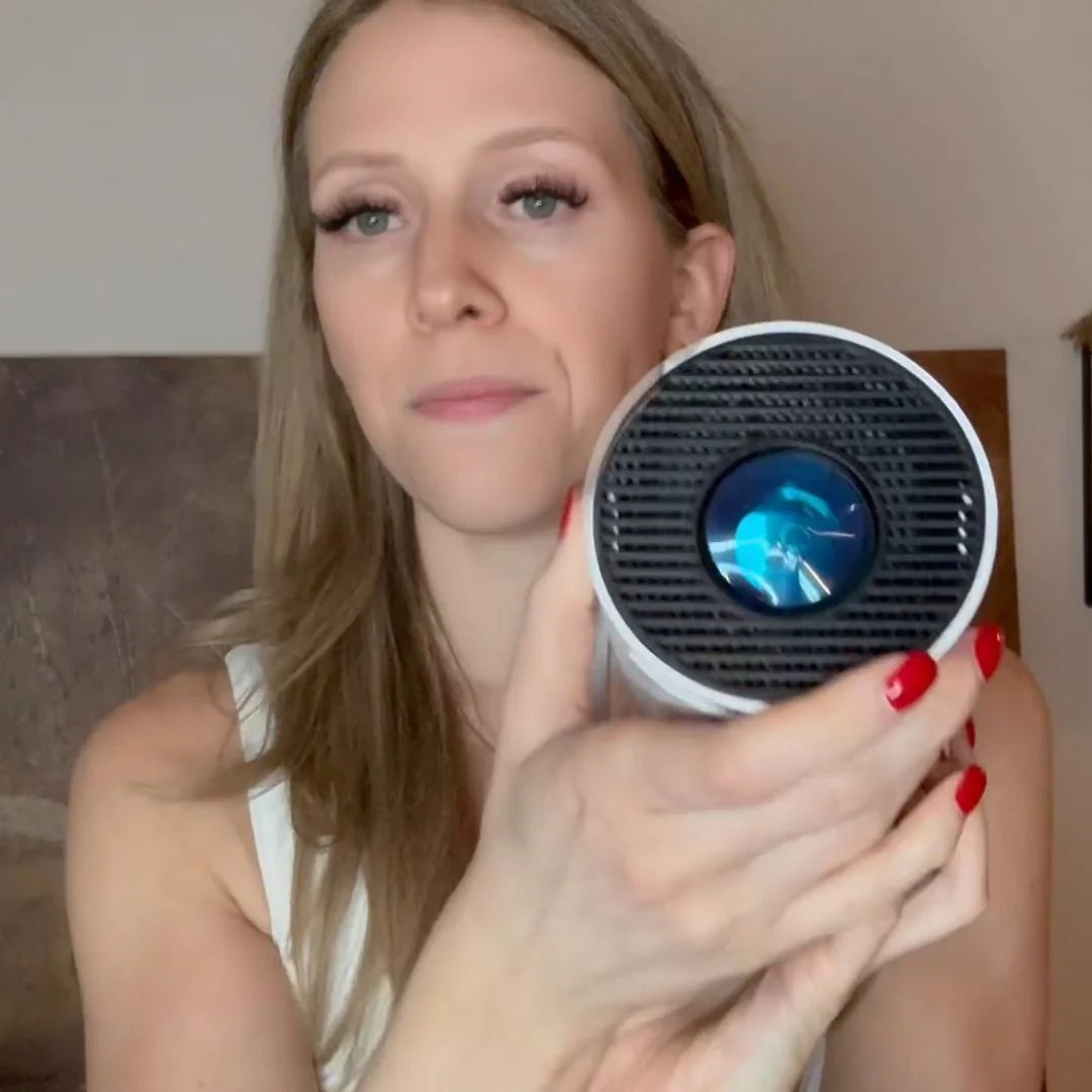 A woman with blonde hair and red nails holds a small, round, white and black projector.