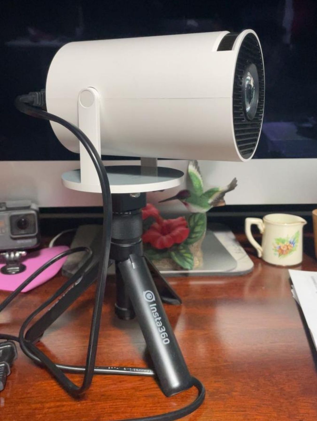 A white, cylindrical projector mounted on a small, black Insta360 tripod on a wooden desk.