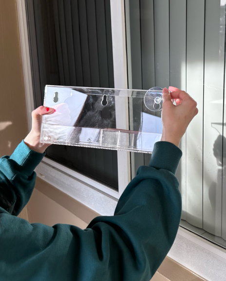 Person holding a clear plastic shelf with suction cups near a window.