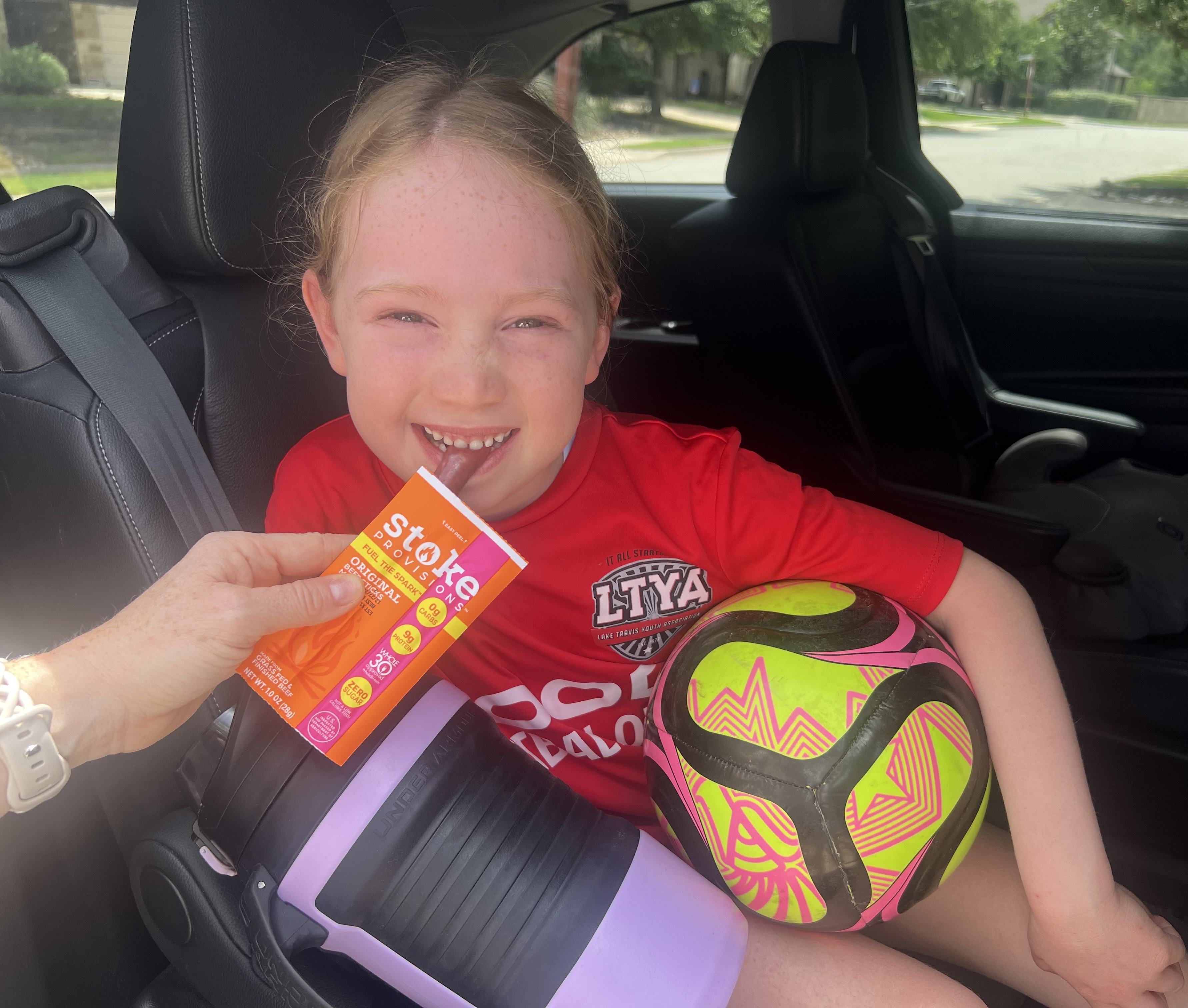 A young girl in a red soccer jersey smiles in a car, holding a soccer ball and a snack.