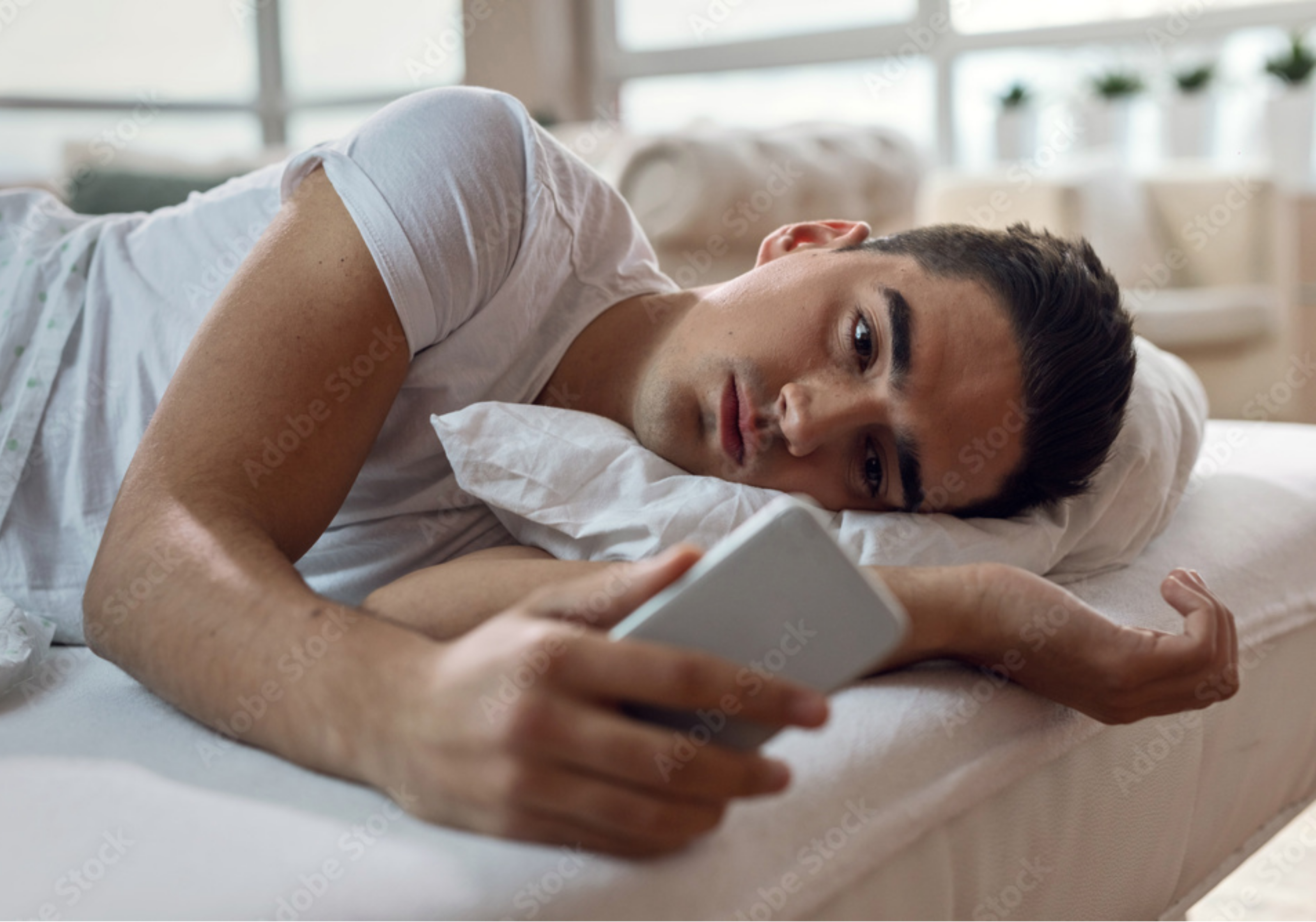 A man lies in bed in a dark room, looking at his smartphone.