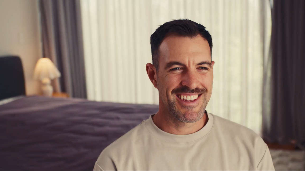Smiling person sits in a bedroom with curtains and a lamp.