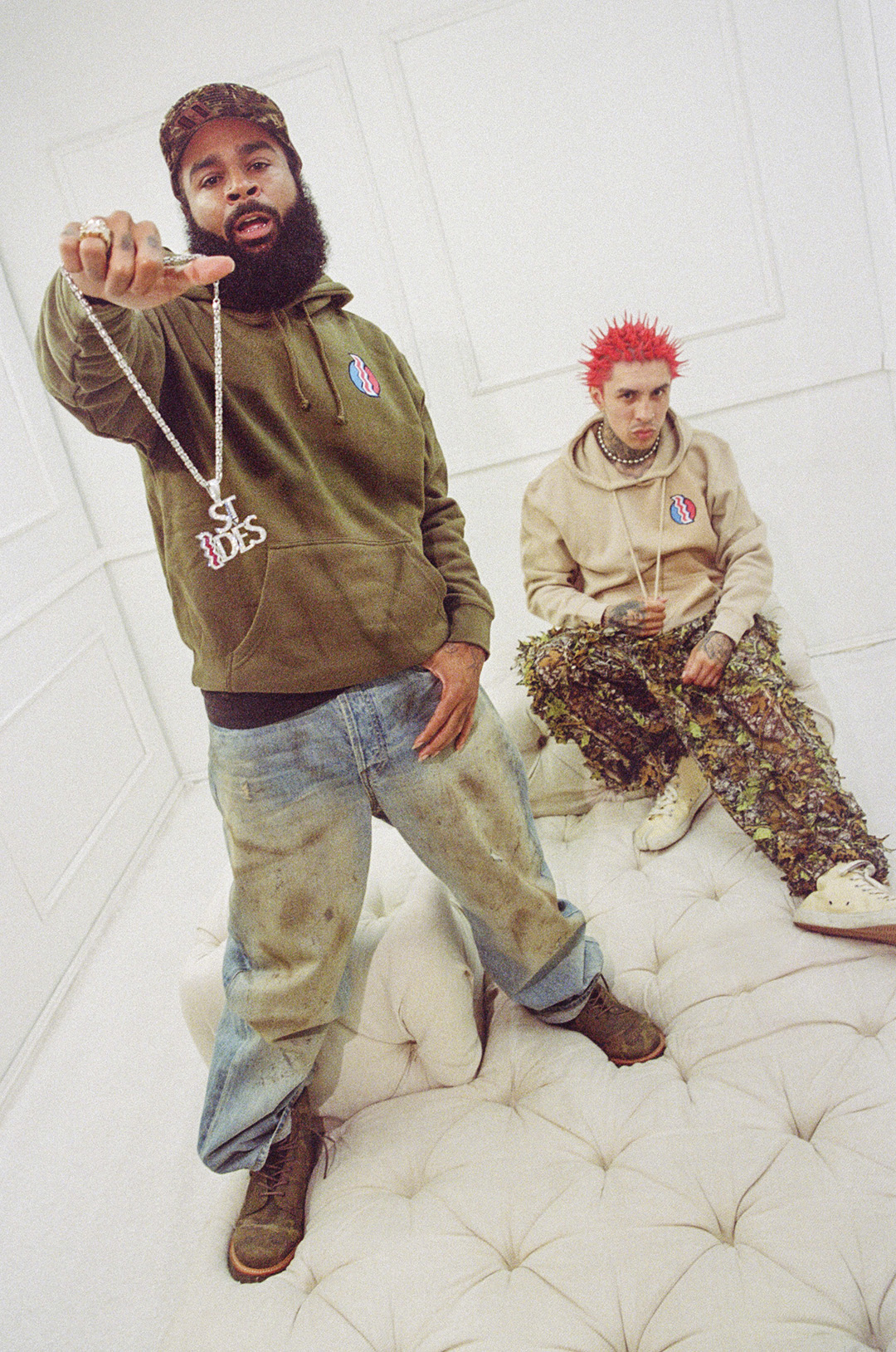 Two men posing in a white room, one holding out a large chain necklace towards the camera.