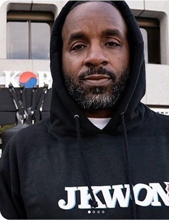Person in a black hoodie stands outdoors in front of a building.