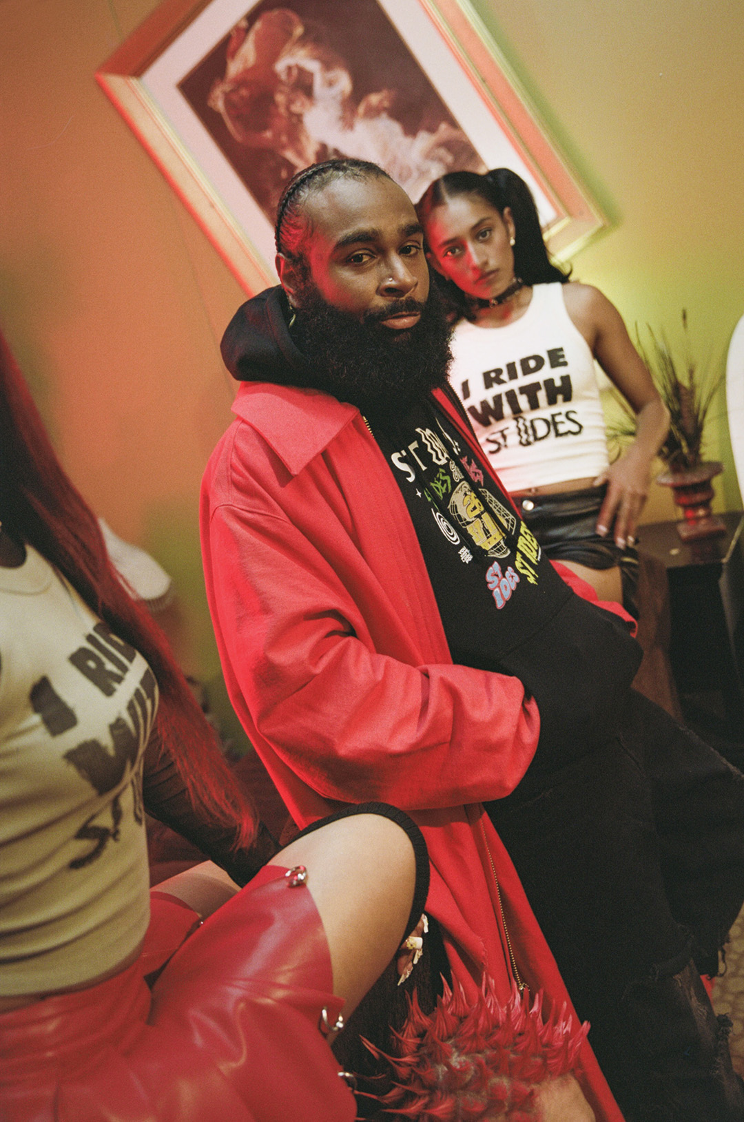 A bearded man in a red jacket sits and looks at the camera, with two women behind him.