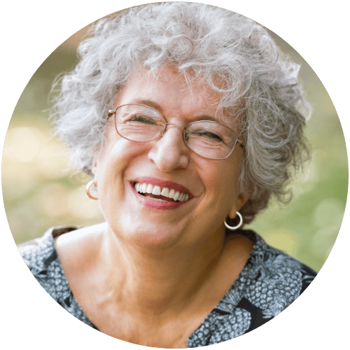 Smiling person with curly hair and glasses, wearing a patterned top, outdoors.