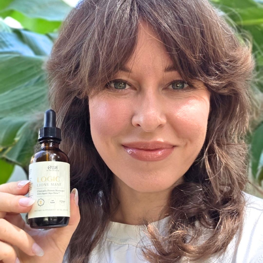 A smiling woman with brown hair holds a small dropper bottle of Logic Lions Mane dietary supplement.