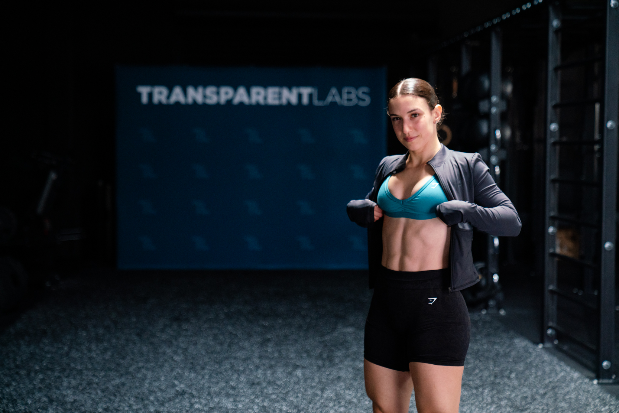 A fit woman in a gym opens her jacket to show her abs in front of a Transparent Labs sign.