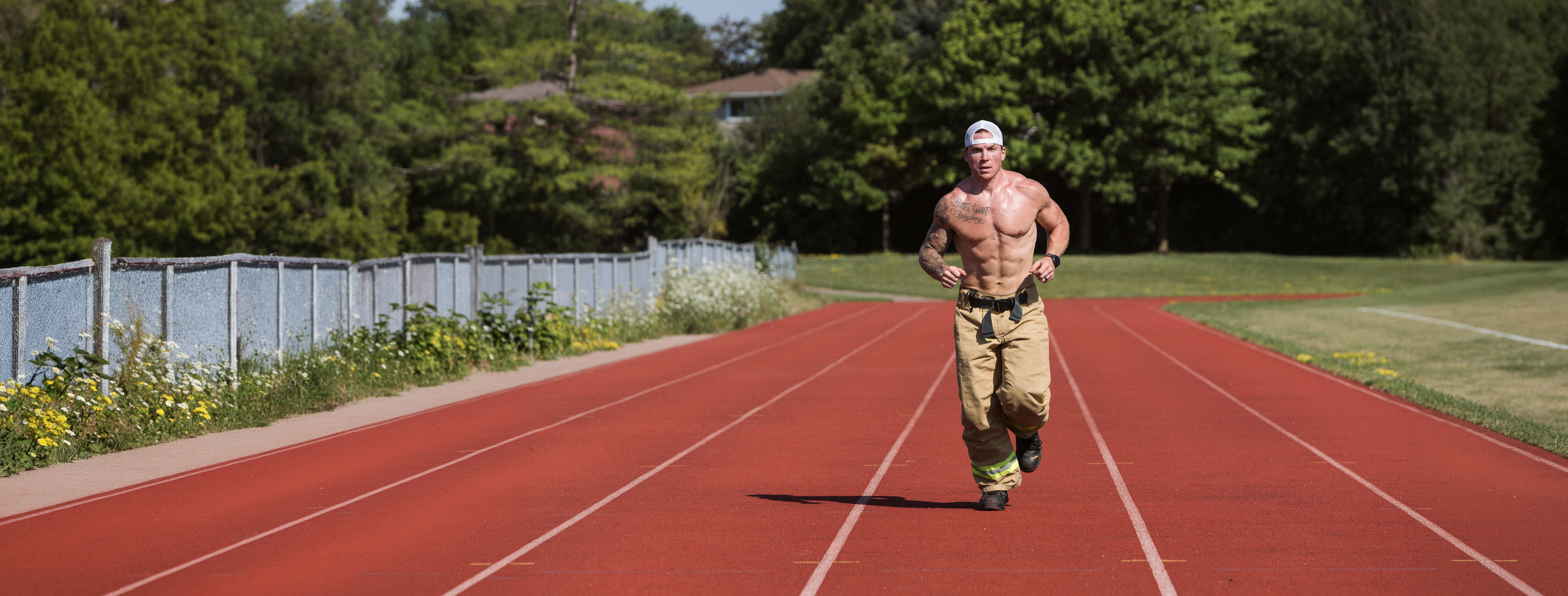 A shirtless, muscular man wearing firefighter pants runs toward the camera on a red running track.