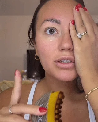 A woman with red nails covers one eye to show her styled eyebrow while holding a product.